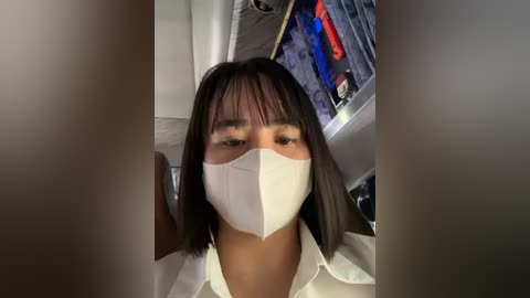 Media: Video of an Asian woman with straight black hair and a white face mask, wearing a white shirt, standing in a cluttered, dimly-lit room with hanging clothes and a partially visible sink.