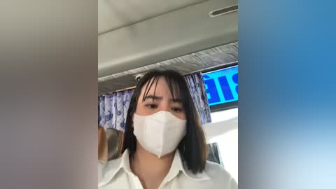 Media: Video of an Asian woman with straight black hair, wearing a white surgical mask, white shirt, and dark pants, standing in a plane with blue and white patterned curtains, and a bright blue LED display behind her.