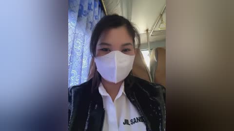 Media: Video of a young woman with light skin and straight, shoulder-length brown hair, wearing a white surgical mask, black jacket, and white shirt with \"Jl. Sangkuriang\" text. Background features a blue curtain with white patterns, and part of a room with a ceiling fan.