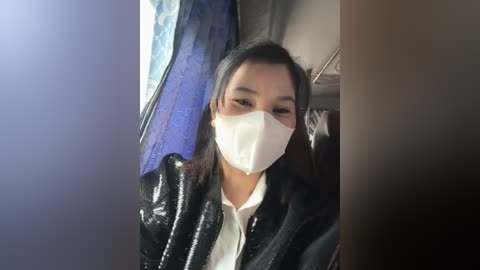 Media: Video of an Asian woman with straight black hair, wearing a white face mask, black leather jacket, and white shirt, in a dimly lit room with blue curtains and a brown wall.