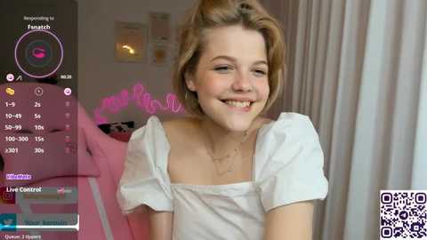 Media: Video of a young, fair-skinned woman with short, blonde hair, smiling, wearing a white off-shoulder top, in a cozy room with pink decor, and a QR code in the bottom right corner.