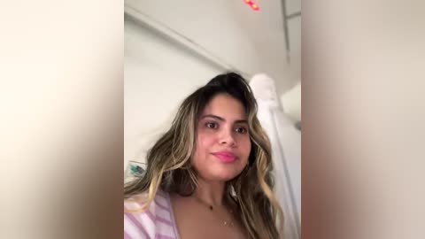 Media: Video of a young Latina woman with long, wavy, dark hair and light skin, wearing a pink and white striped top. She's smiling in a bathroom with white tiles and a shower curtain.