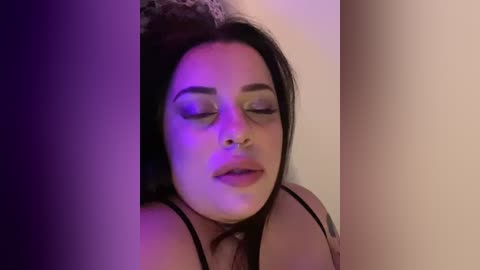 Media: Video of a woman with long black hair, fair skin, and closed eyes, wearing a black bra, in a dimly lit room with purple lighting, leaning against a beige wall.