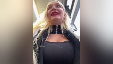 Media: Video of a blonde woman with fair skin, red lipstick, wearing a black jacket over a sheer top, smiling in a dimly lit car.