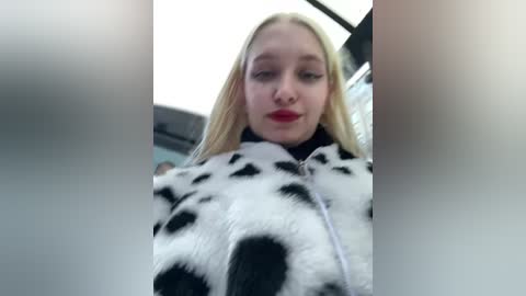 Media: Video of a blonde woman with fair skin, wearing a black and white cow print jacket, sitting in a car with a blurred background.