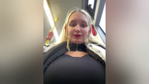 Media: Video of a blonde woman with fair skin and red lipstick, wearing a black dress, standing in a subway car. Background shows fluorescent lights and blurred passengers.