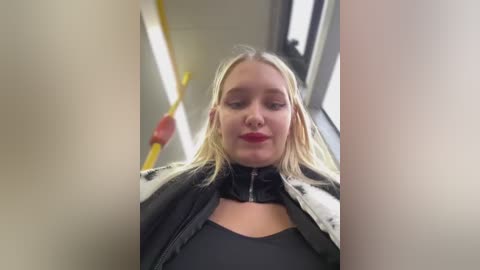 Media: Video of a blonde woman with red lipstick, wearing a black jacket, sitting on a bus with yellow handrails.