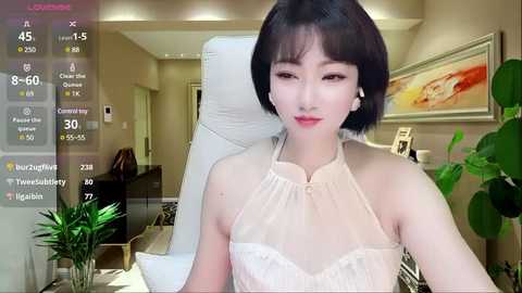 Video of a young East Asian woman with short black hair and pale skin, wearing a sheer white lace top, seated in a modern living room.