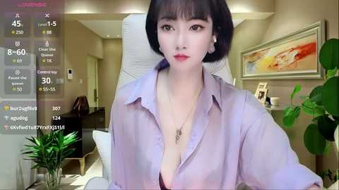 Video of an East Asian woman with short black hair and light skin, wearing a loose lavender blouse, sitting in a modern living room with beige walls and abstract art.