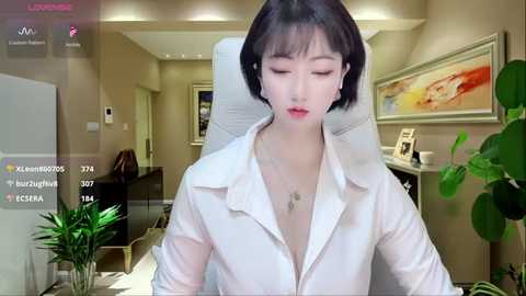 Media: Video of a young East Asian woman with fair skin and short black hair, wearing a white shirt, sitting in an office with a framed abstract painting on the wall.