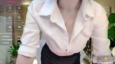 Media: Video of a fair-skinned woman wearing a white, button-down shirt and black skirt, taken from a low angle. Background includes a calendar, a plant, and a blurred, modern room.