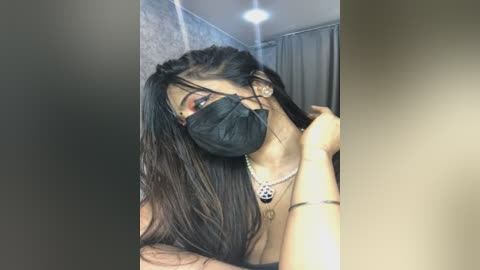 Media: Video of a young woman with long black hair, wearing a black face mask, gold necklace, and a grey top, standing indoors with a grey wall and curtains in the background.