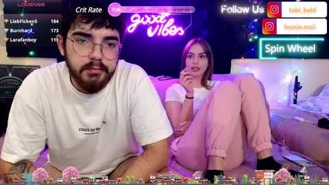 Media: Video of a young man with glasses and beard, wearing a white t-shirt, sitting next to a young woman with long hair, wearing a pink top and pants, in a dimly-lit, neon-lit room with \"good vibes\" and \"spin wheel\" graphics.