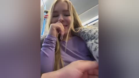 Media: Video of a blonde woman with long hair, wearing a lavender sweater and a fur-trimmed jacket, yawning while seated in a train. The background features blue and white interior elements.