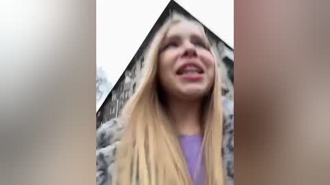 Media: A blurry video of a smiling, blonde-haired woman wearing a purple top and a fur-trimmed coat, standing outdoors against a background of blurred buildings.