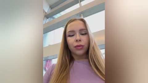 Video of a young Caucasian woman with long blonde hair, wearing a lavender sweater, standing in a modern, brightly-lit room with large windows and white walls.