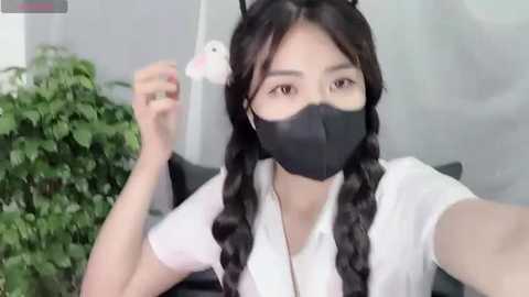 Media: Video of an East Asian woman with long black braids, wearing a white blouse, black face mask, and black cat ears, seated in an office with a green plant in the background.