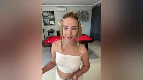 Media: Video of a blonde woman wearing a white crop top and a gold headpiece, standing in a dimly lit room with a pool table and abstract wall art.