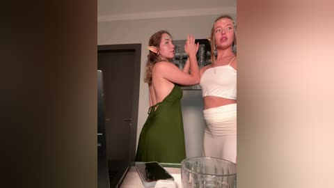 Media: Video of two women in a kitchen: a brunette elf in a green dress and a blonde in white pants and crop top.