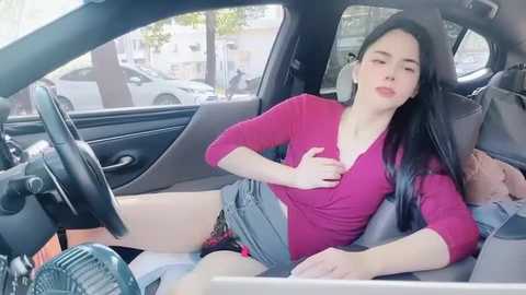 Media: Video of an Asian woman with long black hair, wearing a red long-sleeve shirt and denim shorts, sitting seductively in a car, with her legs spread, eyes closed, and a suggestive expression.