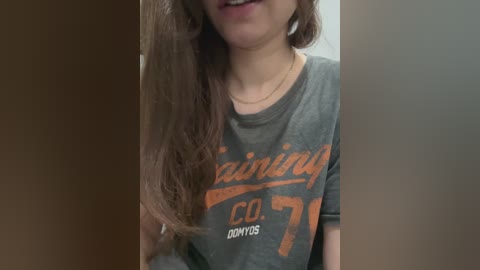 Media: Video of a smiling young woman with long brown hair, wearing a grey T-shirt with orange \"Lining\" and \"Co. 197\" text, standing in a doorway.