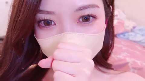 Media: Video of an Asian woman with light skin and long brown hair, wearing a beige face mask, holding it in front of her mouth, with a blurred background featuring a pink bedspread.