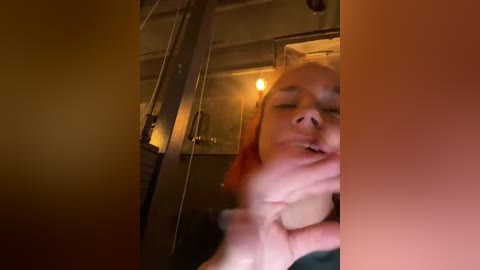 Media: A video of a woman with orange hair, wearing a dark top, looking upward, with her hands near her mouth, in a dimly lit room with warm lighting.