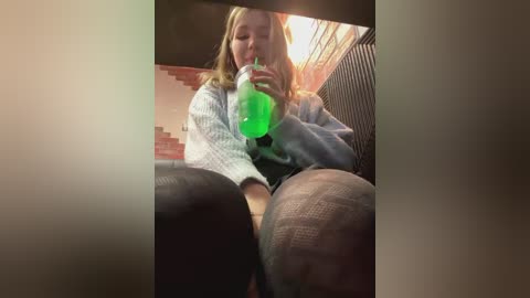 Media: A video captures a young woman with long blonde hair, wearing a light blue sweater and grey patterned leggings, drinking a bright green beverage. She is seated in a dimly lit room with brick walls and a metal chair.