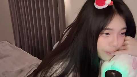 Media: Video of a young East Asian woman with long black hair, wearing a plush red cat headband, lying on a bed, resting her head on a stuffed animal, in a dimly lit room with grey curtains.