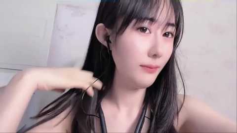 Media: Video of a young Asian woman with long black hair, fair skin, and a delicate facial structure. She's adjusting her hair with one hand, wearing a black top. Background features a white wall and a partially visible drawing.