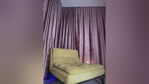 Media: Video of a minimalist room with a yellow armchair against a backdrop of floor-to-ceiling, flowing, light pink curtains. A small, blue light bulb is on the floor near the chair. The room has a neutral color palette.