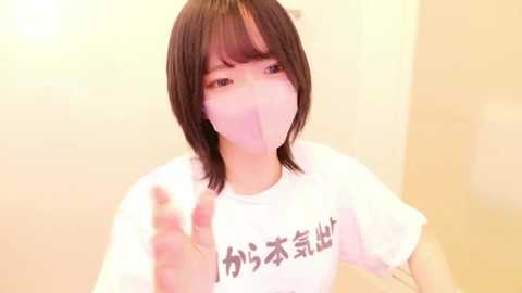 Media: A video of an East Asian woman with short, straight black hair, wearing a white t-shirt and a pink face mask, giving a thumbs-up gesture against a blurred, beige background.