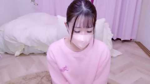 Media: Video of an East Asian woman in a pastel pink room, wearing a pink sweatshirt and face mask, sitting on a beige rug, with a bed in the background.