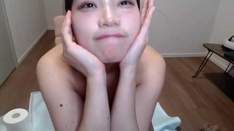 Media: Video of an East Asian woman with pale skin, small breasts, and short black hair, wearing only a towel, resting her head on her hands in a minimalist room with wooden floors and white walls.