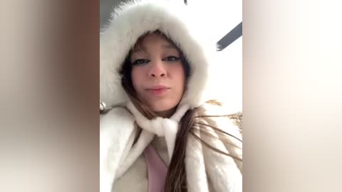 Media: Video of a young woman with light brown skin, wearing a white fur hooded coat, making a pouty face, indoors with a blurred background.