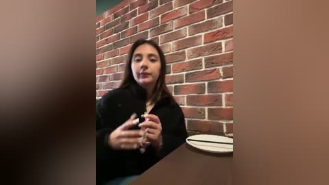 Media: A video of a young woman with straight, dark hair, wearing a black top, gesturing with her hands, seated at a table with a white plate and glass. She is in front of a red brick wall, creating a rustic ambiance.