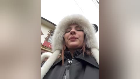 Media: Video of a young Caucasian woman with fair skin, wearing a black jacket with white fur trim, and a white fur hat. She stands in front of a red building with a gray sky and utility wires in the background.