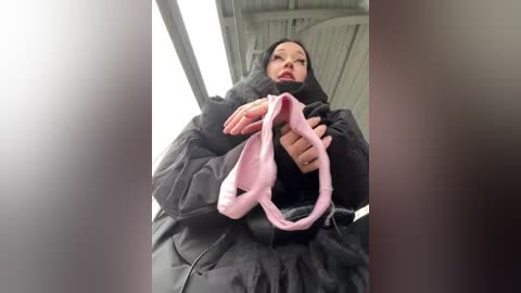 Media: Video of a woman in a black winter coat, holding pink underwear, with a blurred background featuring a metal structure. She has a neutral expression.