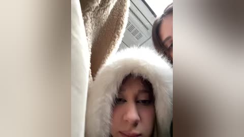 Media: Video of a young woman with fair skin and long brown hair, wearing a fluffy white and brown fur hood, looking down. Background shows blurred faces and a beige blanket.