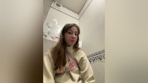 Media: Video of a young woman with long brown hair, wearing a beige sweater and a pink bra, in a small, dimly lit bathroom with white tiled walls and a decorative blue border.