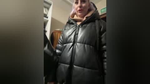 Media: A video shows a person wearing a black puffy jacket with a light tan hoodie underneath, standing in a dimly lit room with beige walls and a wooden chair.