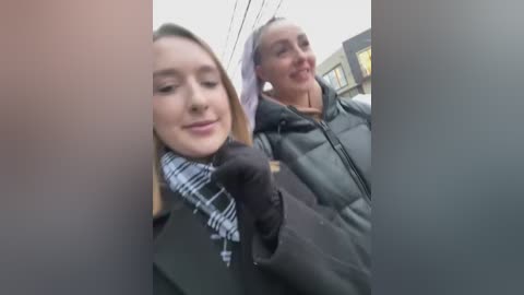 Media: Video of two women, one with long blonde hair and a black scarf, the other with short brown hair, both wearing winter coats, standing on a city street.