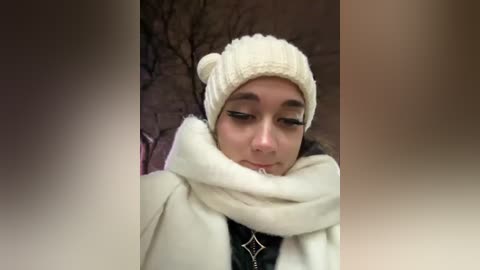 Media: Video of a young woman with fair skin and dark hair, wearing a cream beanie and white scarf, standing in a blurred, wintry background with bare trees.