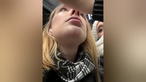 Media: A close-up video of a blonde woman with fair skin, wearing a black coat and a plaid scarf, wiping a tear from her eye, taken in a public setting.