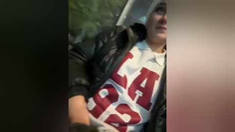 Media: A video of a person wearing a black jacket over a white polo shirt with large red letters, sitting in a car with blurred outdoor background.