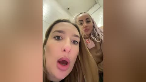 Media: Video of two women with fair skin and light brown hair, one wearing a beige top, the other in a pink blouse, both with shocked expressions.