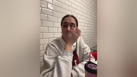 Media: Video of a woman with brown hair, wearing a white hoodie, blowing bubbles in a bathroom with white tiled walls and a pink cup.