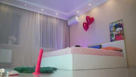 Media: Video of a modern bedroom with beige walls, a white bed, red heart balloons, and a pink candle on the floor.