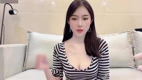 Media: Video of an Asian woman with long, straight black hair, wearing a low-cut, black and white striped top, sitting on a light-colored couch with white pillows. She holds a phone, possibly filming herself. Background includes a lamp and a neutral-colored wall.