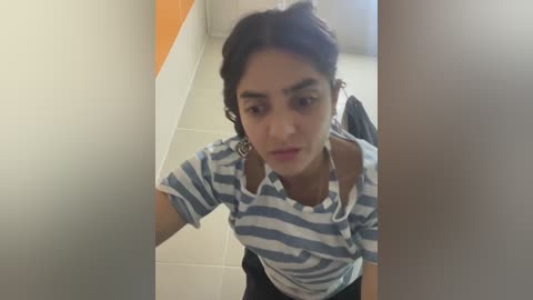 Media: Video of a young woman with medium-dark skin, wearing a white and blue striped polo shirt, taking a selfie in a bathroom with tiled walls.